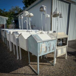 A row of weathered, metal meteorological and environmental monitoring stations with attached sensors, lined up outdoors beside a warehouse, representing industrial and science surplus donations for GiveSurplus.org.