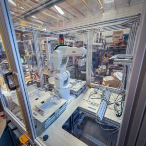 A white ABB robotic arm enclosed in a transparent safety case within a workshop filled with tools and materials, part of GiveSurplus.org’s industrial and science surplus donations initiative.