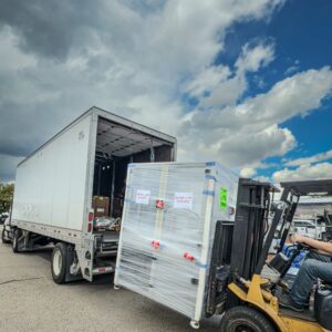 A forklift unloading a large, plastic-wrapped crate labeled “Do Not Lift This Side” onto a semi-truck under a partly cloudy sky, depicting industrial and science surplus being shipped to GiveSurplus.org donations.